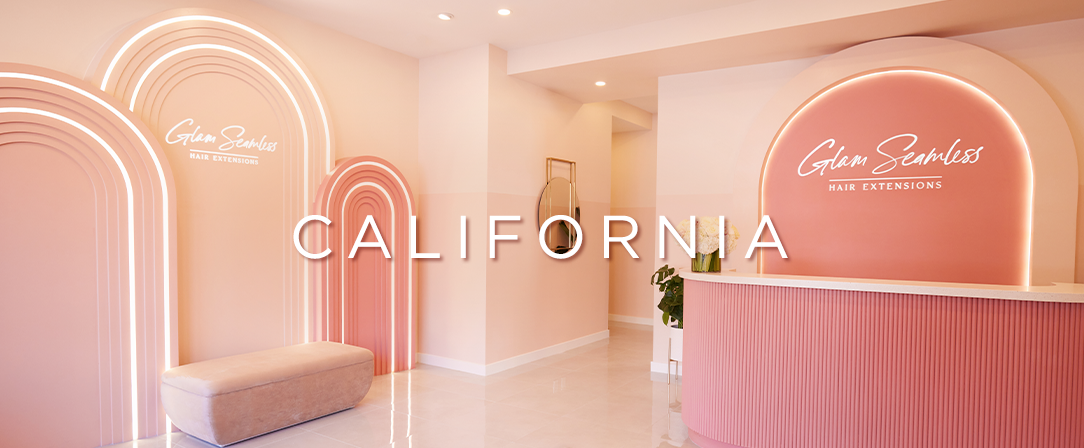 A Glam Seamless Hair Studio with Pink walls, Mirrors, fresh flowers, a waiting sofa, and a reception desk with California text.