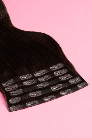 A set of dark brown clip-in hair extensions with secure black clips, laid on a pink background.