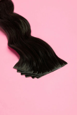 A black tape-in hair extension with a wavy texture is displayed against a pink background.