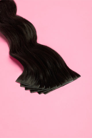 A natural black hair extension is displayed on a soft pink background, showcasing its sleek and elegant design.