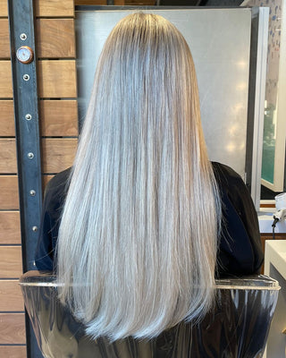 Woman showcasing Glam Seamless rooted platinum blonde hair extensions in a salon and sitting on a chair.