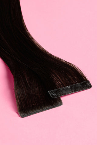 A pair of premium Tape natural black hair extensions are displayed on a soft pink background.