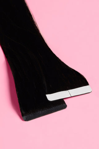 A pair of premium remy tape-in jet black hair extensions is displayed on a pink surface.