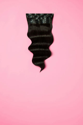 A black clip-in hair extension is displayed prominently against a soft pink background.