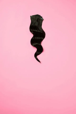 A single black wavy hair extension clip-in piece displayed against a pink background.