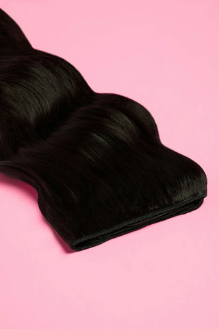 Black wavy hair extensions stacked on a pink background, highlighting shine, thickness, and smooth texture.